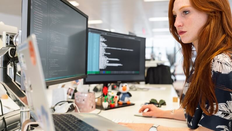Red head woman coding with two monitors and a laptop in front of her at a desk.